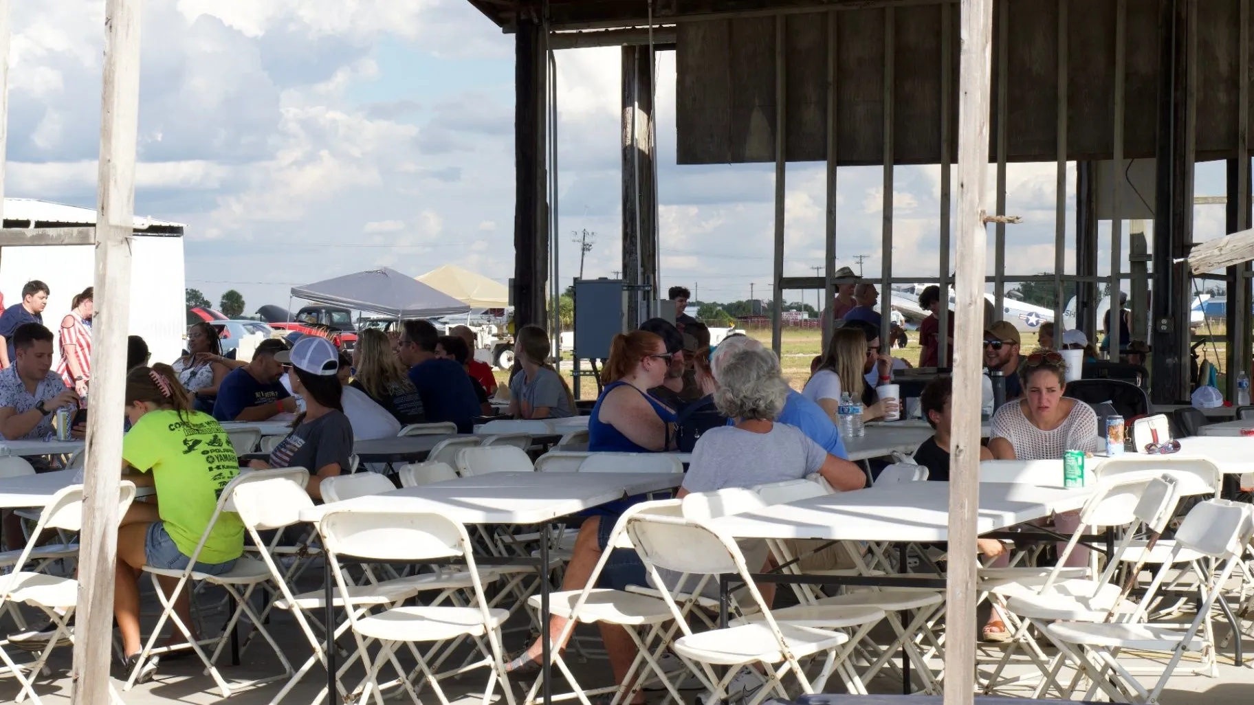 People sit together at long tables beneath a covered outdoor structure.