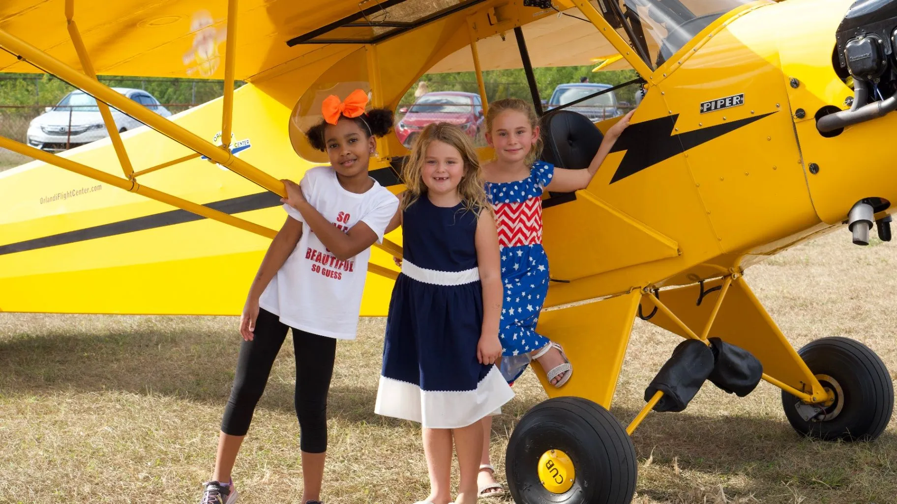 Three children smile beside a bright yellow airplane at an outdoor aviation event.