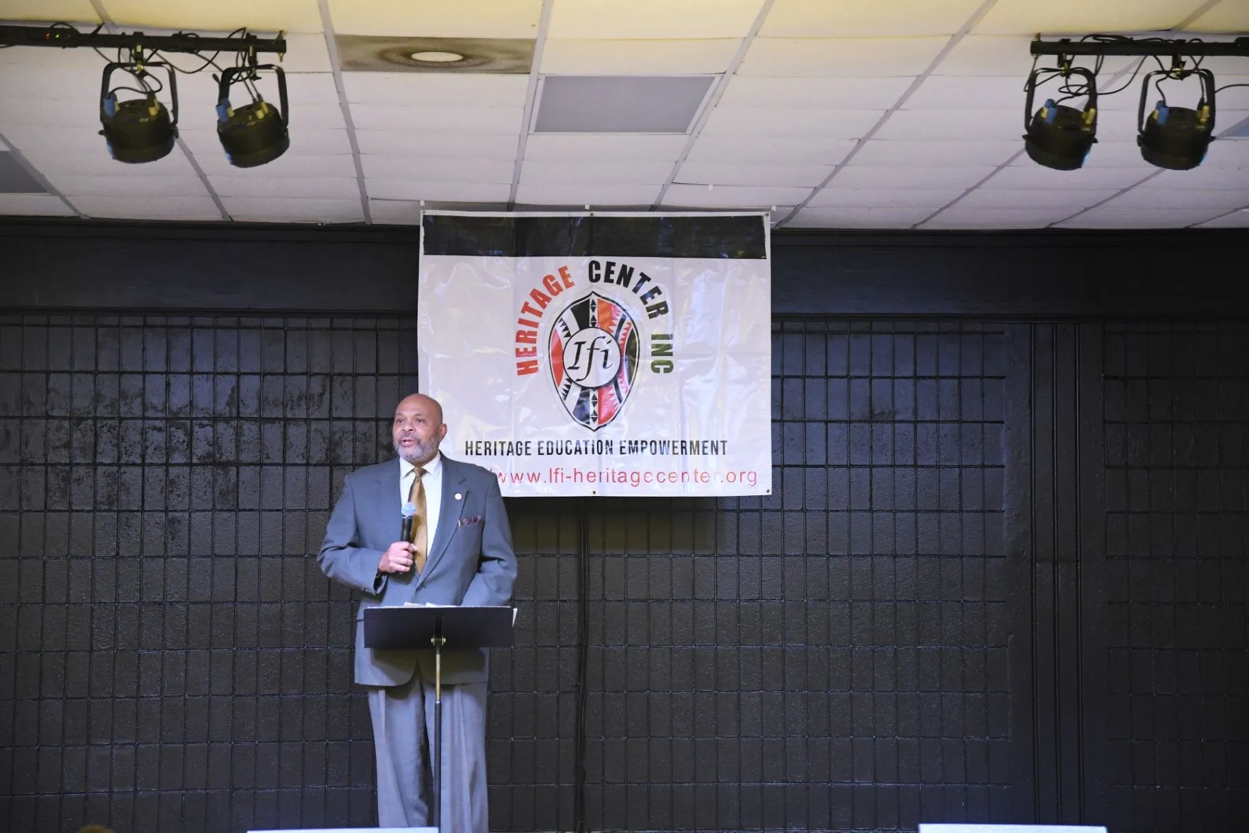 A speaker presents on a stage beneath an Ifi Heritage Center banner.