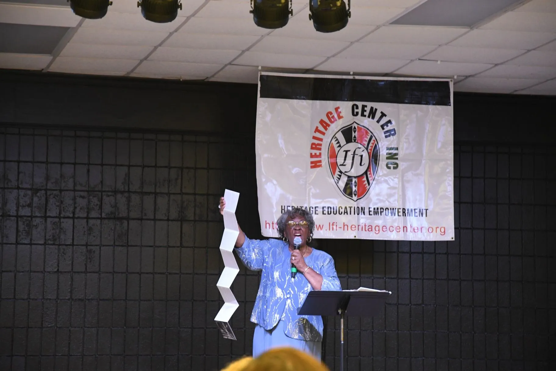 Ifi Heritage Center speaker presenting on stage with a microphone and a paper in hand beneath the Heritage Center banner.