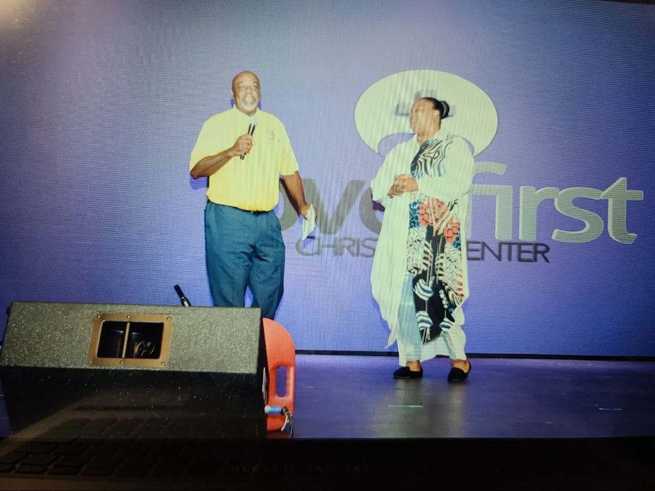 Two community members on a stage, one speaking into a microphone and one wearing a patterned outfit.
