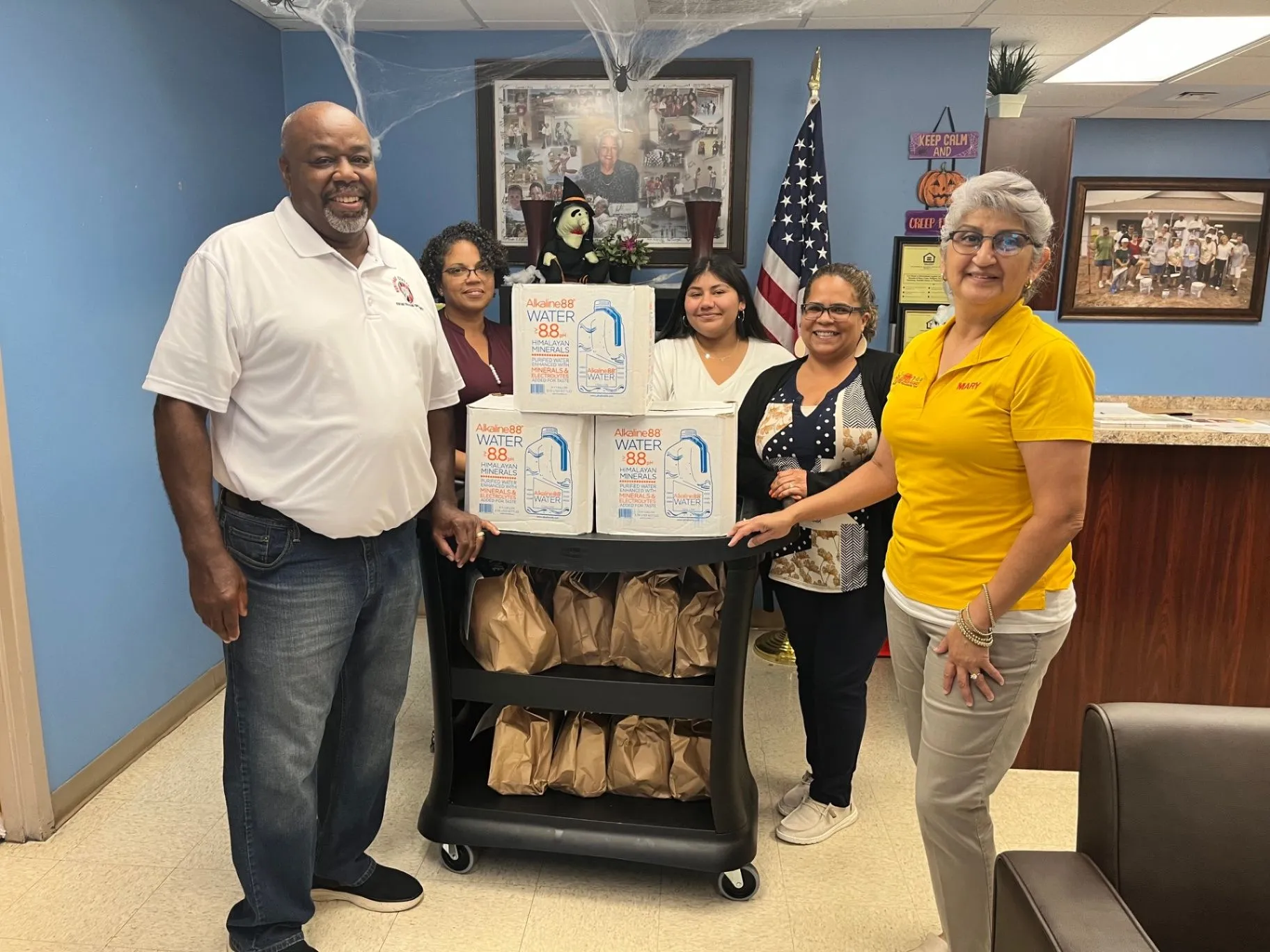 Five people pose around a cart stacked with boxes of water inside a community office.