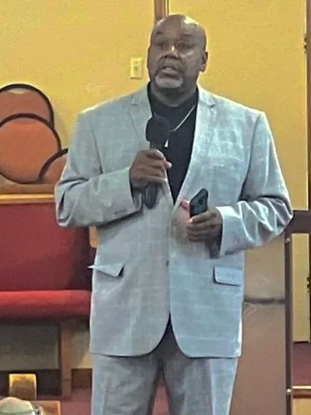 A man in a light suit holds a microphone while speaking to a gathering.