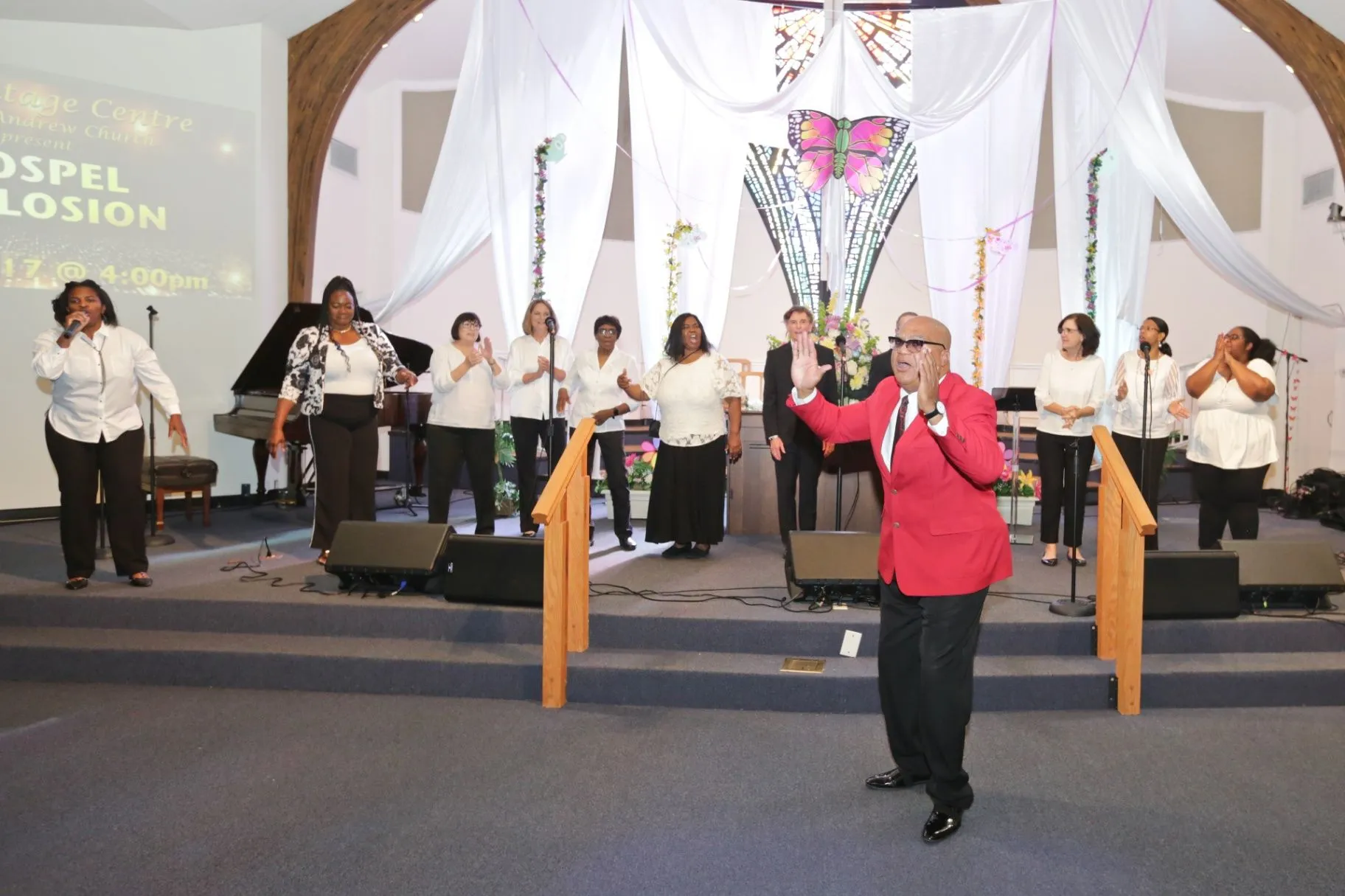 A worship and performance group sings on a decorated church stage while a man in a red jacket gestures in front.