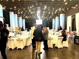 People gather at tables beneath string lights inside a decorated event space.