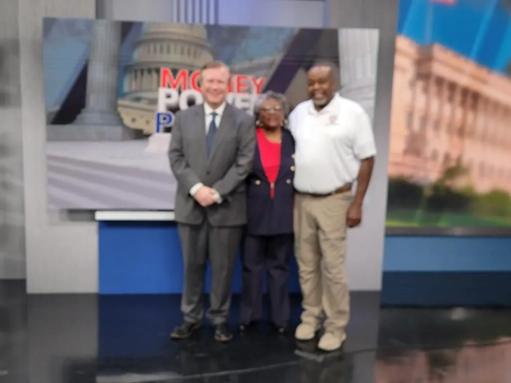 Three people stand together in a studio or public media setting.