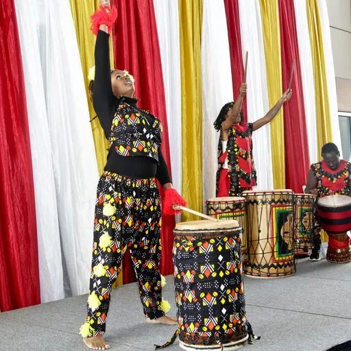 Performers in colorful patterned clothing play drums in front of red, white, and gold curtains.